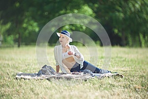 A woman in a hat is holding a glass of red wine in her hand at a picnic. Lady sitting on the grass with a blanket