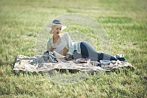 A woman in a hat is holding a glass of red wine in her hand at a picnic. Lady sitting on the grass with a blanket