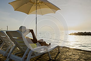 A woman in a hat is lying on a deckchair on the beach at dusk