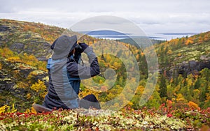 A woman in a hat on a mountain slope looks through binoculars in autumn against the backdrop of a lake and mountains