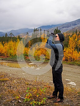 A woman in a hat on a mountain slope looks through binoculars in autumn against the backdrop of a lake and mountains