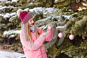 A woman in a hat and pink down jacket hangs Christmas balls on a Christmas tree