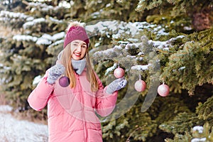 A woman in a hat and pink down jacket hangs Christmas balls on a Christmas tree