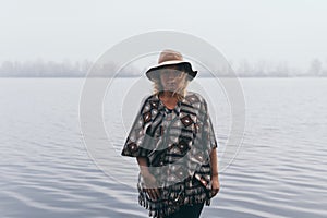 Woman in hat and poncho standing on the river bank and looking towards autumn forest covered with morning fog