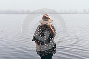 Woman in hat and poncho standing on the river bank and looking towards autumn forest covered with morning fog