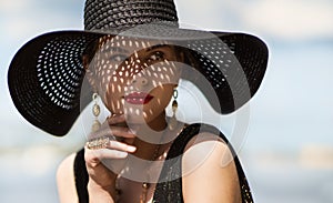 Woman in Hat Portrait. Fashion Luxury Model in Black Summer Hat with Make up and Golden Jewelry. Close up Beauty Face over Sky