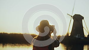 Woman in hat sits on sunset countryside lake quay. Girl takes an old windmill photo using her smartphone. 4K back view.