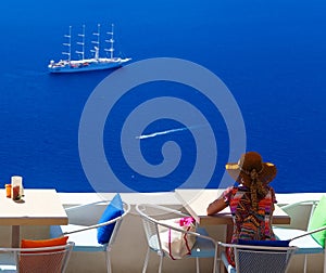 Woman in hat sitting in cafe and enjoys views of sailing cruise ship, Santorini, Fira, Greece