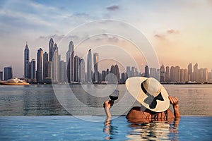 A woman with a hat stands at the edge of a pool and enjoys the view to the Dubai Marina