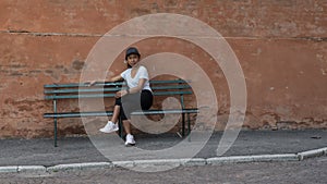 Woman in hat, tshirt, black pants and sneakers sitting on park bench with legs crossed with another park bench to her right
