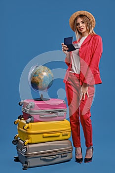 Woman in hat, white blouse, red pantsuit, black heels. She is holding passport and ticket, posing on blue background