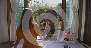 Woman having breakfast inside bungalow and treats elephant from the balcony