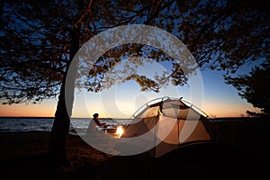 Woman having a rest at night camping near tourist tent, campfire on sea shore under starry sky