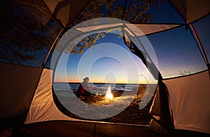 Woman having a rest at night camping near tourist tent, campfire on sea shore under starry sky