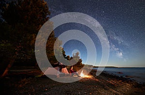 Woman having a rest at night camping near tourist tent, campfire on sea shore under starry sky