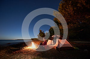 Woman having a rest at night camping near tourist tent, campfire on sea shore under starry sky