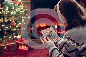 Woman in headphones with christmas gift box in hand sitting on fluffy plaid near fireplace and christmas tree.