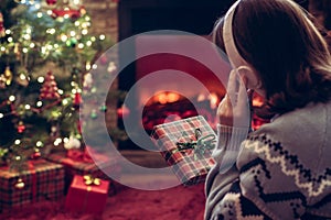 Woman in headphones with christmas gift box in hand sitting on fluffy plaid near fireplace and christmas tree.