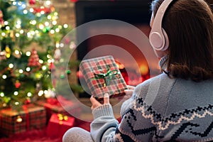 Woman in headphones with christmas gift box in hand sitting on fluffy plaid near fireplace and christmas tree.