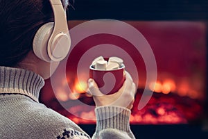 Woman in headphones with cup of hot cocoa and marshmallow sitting and warming at winter near fireplace flame