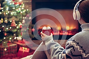 Woman in headphones with cup of hot cocoa and marshmallow sitting and warming at winter near fireplace flame and christmas tree
