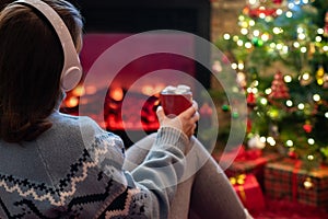 Woman in headphones with cup of hot cocoa and marshmallow sitting and warming at winter near fireplace flame and christmas tree