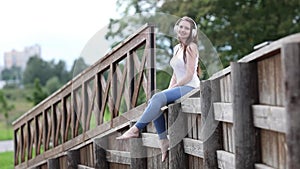 Woman with headphones dangles her legs while sitting on fence in public park.