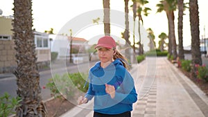 Woman with headphones in ears runs down the street along the palm avenue at sunset. Healthy active lifestyle