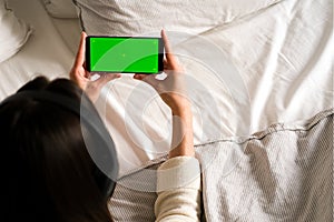 A woman with headphones lies and holds a black mobile phone with a green screen upright