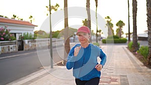 Woman with headphones runs down the street along the palm avenue at sunset. Healthy active lifestyle. Slow motion