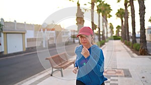 Woman with headphones runs down the street along the palm avenue at sunset. Healthy active lifestyle. Slow motion