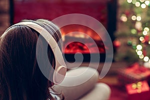 Woman in headphones sitting and warming at winter evening near fireplace flame and  christmas tree.