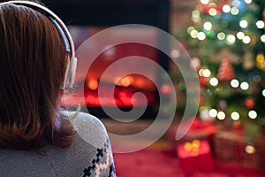 Woman in headphones sitting and warming at winter evening near fireplace flame and  christmas tree.