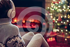 Woman in headphones sitting and warming at winter evening near fireplace flame and  christmas tree.