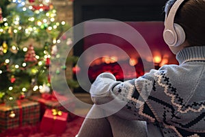 Woman in headphones sitting and warming at winter evening near fireplace flame and  christmas tree.