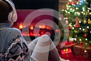 Woman in headphones sitting and warming at winter evening near fireplace flame and  christmas tree.