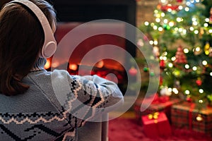 Woman in headphones sitting and warming at winter evening near fireplace flame and  christmas tree.