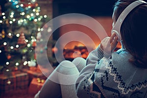 Woman in headphones sitting and warming at winter evening near fireplace flame and  christmas tree.