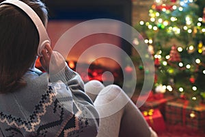 Woman in headphones sitting and warming at winter evening near fireplace flame and  christmas tree.