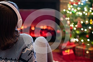 Woman in headphones sitting and warming at winter evening near fireplace flame and  christmas tree.