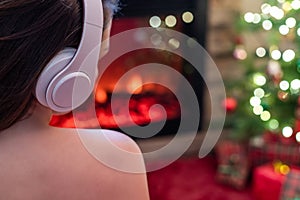 Woman in headphones sitting and warming at winter evening near fireplace flame and  christmas tree.