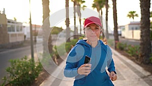 Woman with headphones and smartphone runs down the street along the palm avenue at sunset. Healthy active lifestyle