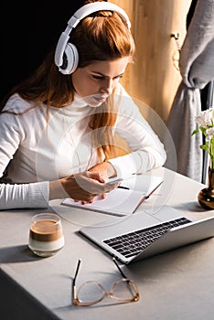 Woman in headphones writing in notepad while watching webinar on laptop in cafe with glass of coffee