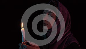 Woman in a headscarf with a burning candle in hands praying to god in darkness