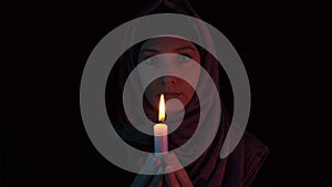 Woman in a headscarf with a burning candle in hands praying to god in darkness, the candle extinguished mystically