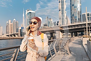 Woman in a headscarf drinking coffee and talks and gossips on the phone with a friend on the Dubai Walk Canal