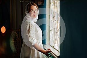 a woman in headscarf and light-colored robes by window in church.