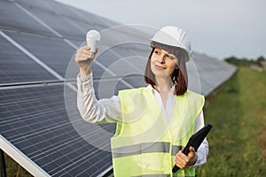 Woman in helmet standing at solar farm and holding bulb