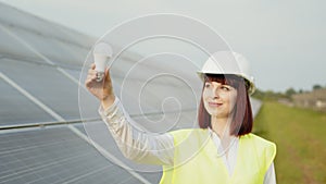 Woman in helmet standing at solar farm and holding bulb. Woman in uniform and white helmet standing at solar farm