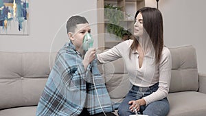 Woman helping illness boy with nebulizer inhalation at home. A young mother helping her son to inhale with a nebulizer at home